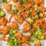 Overhead view of sheet pan General Tso’s chicken with crispy chicken pieces, broccoli florets, and red bell peppers tossed in a glossy sauce and sprinkled with sesame seeds and green onions on foil.