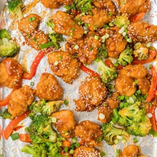 Overhead view of sheet pan General Tso’s chicken with crispy chicken pieces, broccoli florets, and red bell peppers tossed in a glossy sauce and sprinkled with sesame seeds and green onions on foil.