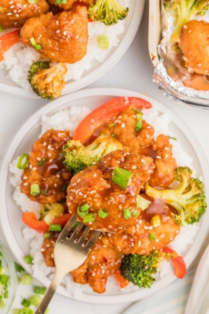 Close-up of General Tso’s chicken over rice with a fork lifting a glazed piece, showing broccoli and red peppers with sesame seeds and green onion garnish.