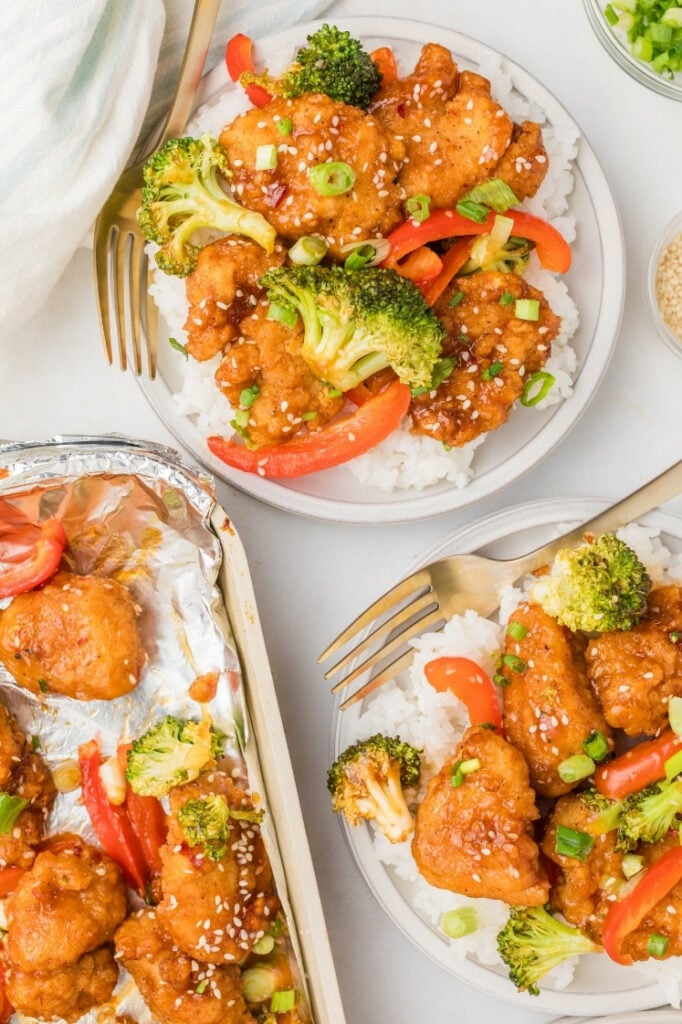 Overhead view of two bowls of General Tso’s chicken over rice with broccoli and red peppers, with sesame seeds and green onions, alongside a sheet pan of extra portions.