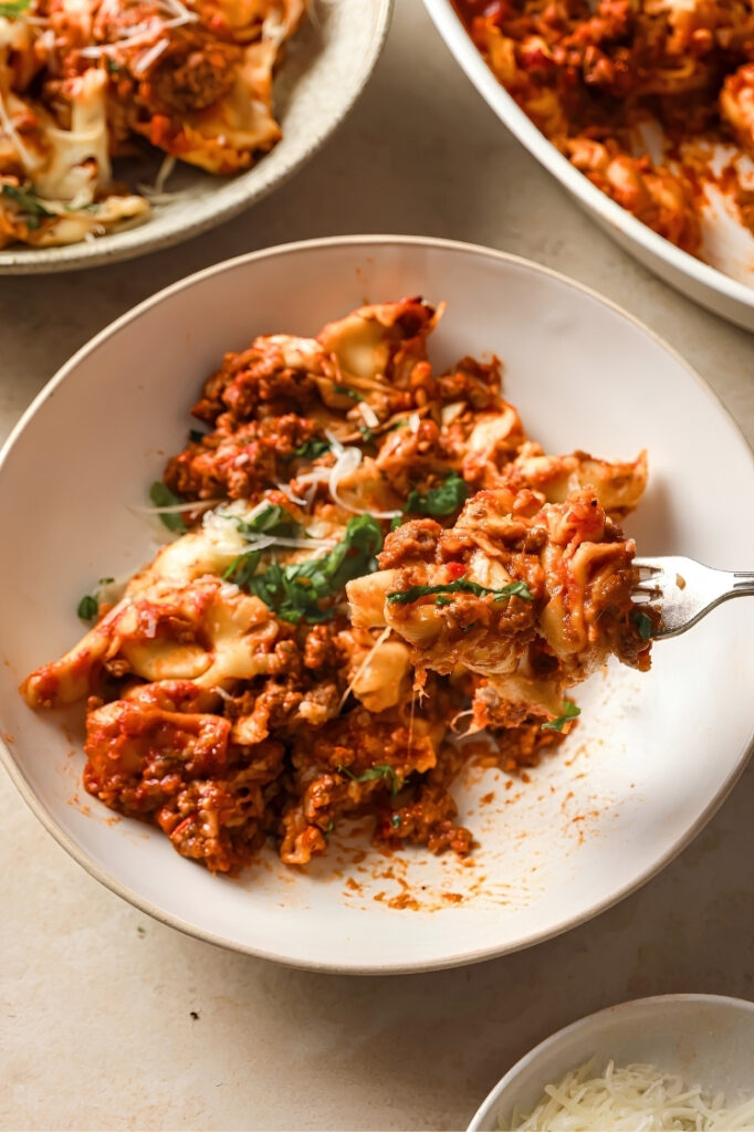Forkful of cheesy tortellini and meat sauce lifted from a plated serving with melted cheese strands visible.