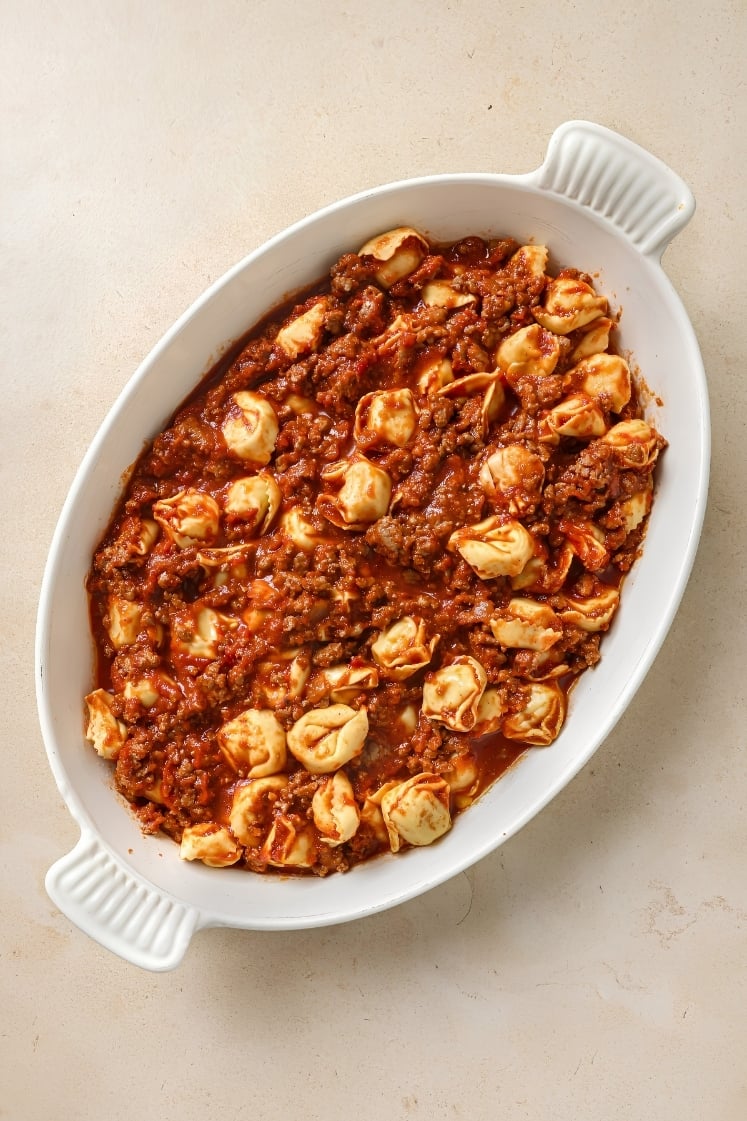 Overhead shot of tortellini coated in hearty meat sauce in a white baking dish before cheese is added.