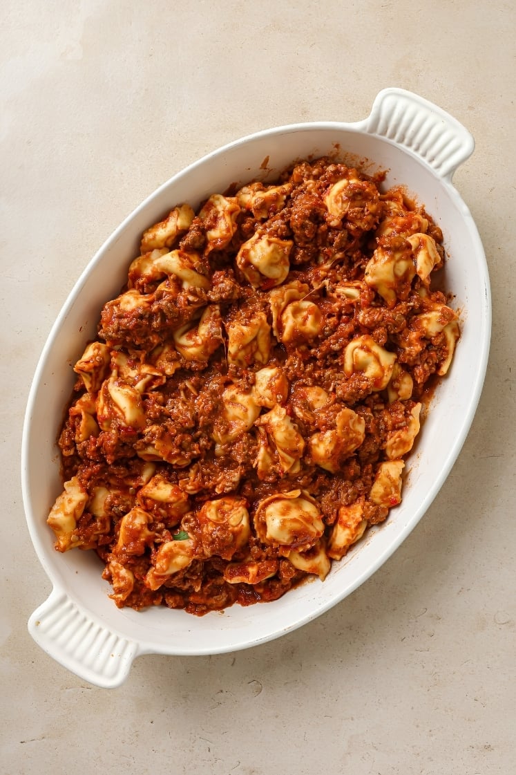 Top view of tortellini and meat sauce spread evenly in a white casserole dish with extra sauce visible around the edges.