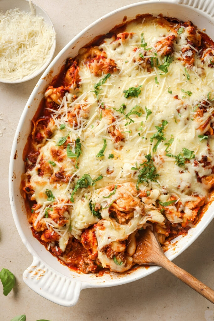 Close overhead shot of baked tortellini being scooped from the casserole dish with melted cheese, basil, and meat sauce visible.