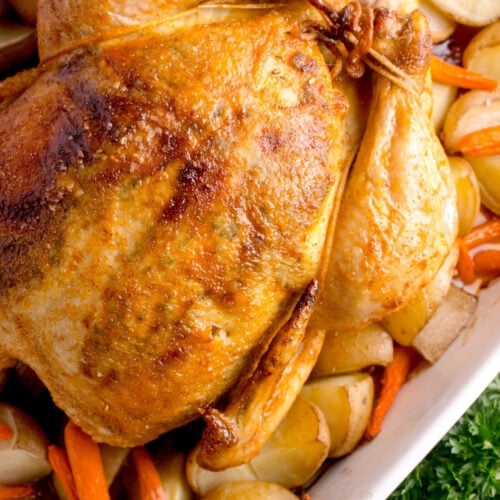 Close-up view of the crispy, glistening golden-brown skin of a roasted whole chicken inside a white baking dish surrounded by carrots and potatoes.