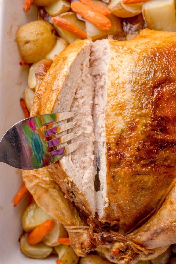 A metal fork piercing a thick slice of juicy, white-meat chicken breast being carved directly from the roasted whole chicken.