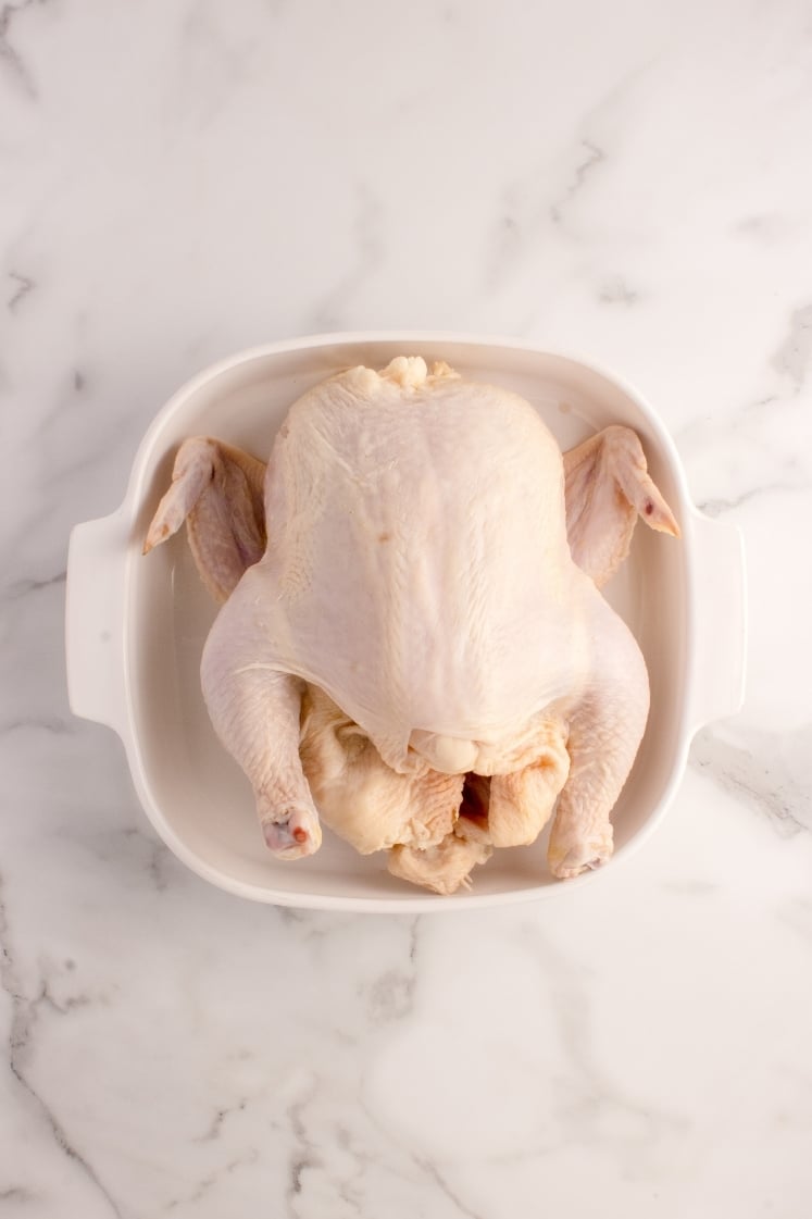 Overhead shot of a whole raw chicken, patted dry, sitting centered inside a white square ceramic baking dish on a marble countertop.
