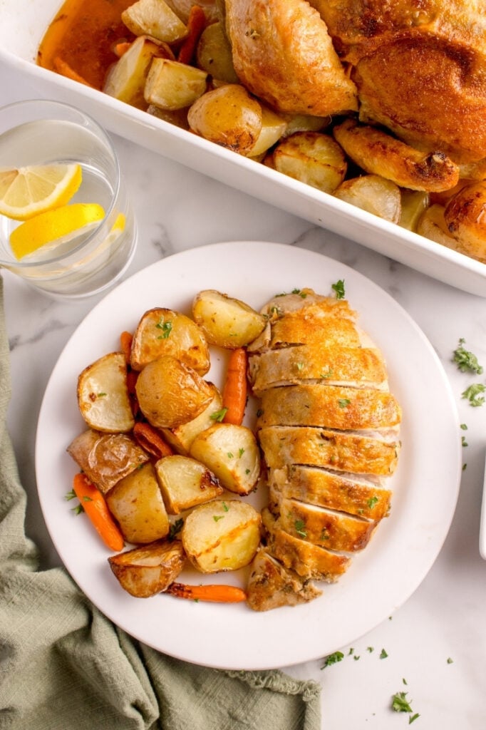 Vertical close-up of a white dinner plate featuring a sliced juicy roasted chicken breast alongside seasoned roasted potatoes and carrots. A baking dish with the rest of the chicken and a glass of lemon water sit in the background.