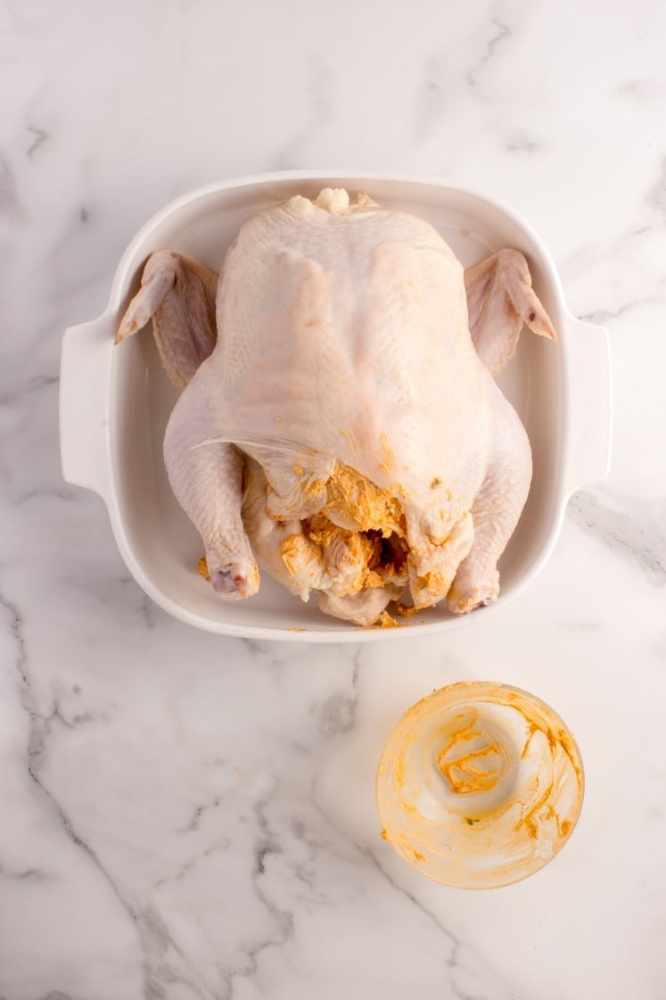 Whole raw chicken inside a white baking dish with seasoned compound butter stuffed generously under the breast skin. An empty glass bowl sits nearby.