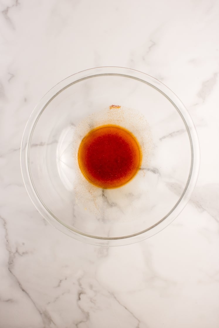 Top-down view of a clear glass bowl containing a bright orange and red melted butter glaze mixed with spices.