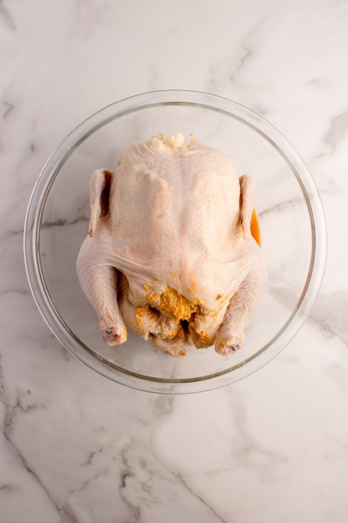 Whole raw chicken sitting inside a large glass mixing bowl, getting prepared to be coated in the spice glaze.
