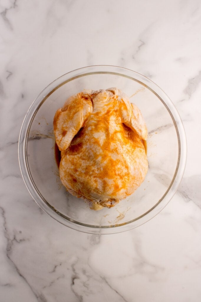 Raw chicken inside a glass bowl completely coated in a vibrant orange spice marinade, with glaze brushed thoroughly over the skin and wings.