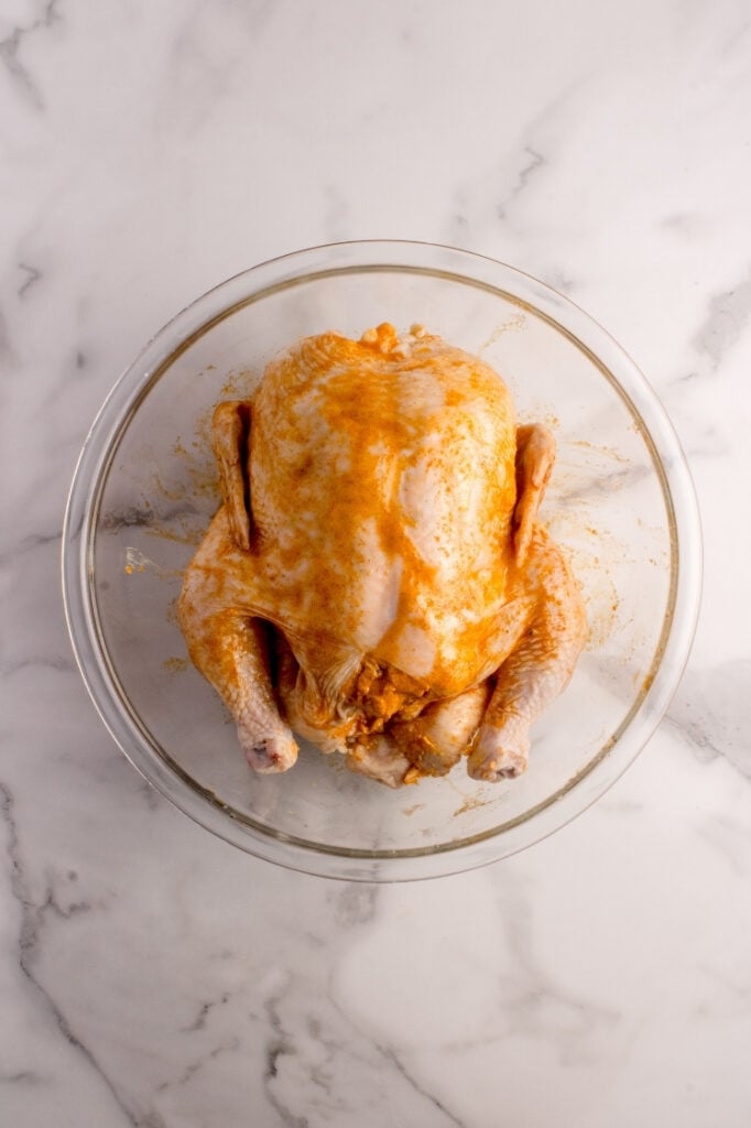 Front view of the marinated whole raw chicken sitting in the glass bowl, showing the cavity packed with seasoning.