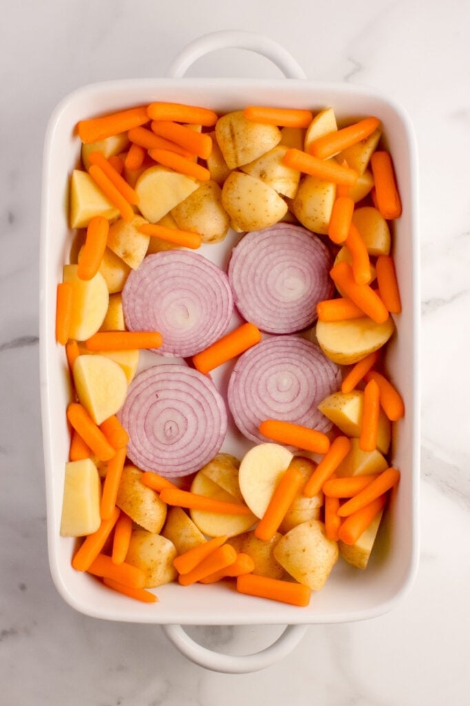A white rectangular baking dish filled with a layer of chopped raw potatoes, baby carrots, and thick round slices of red onion.