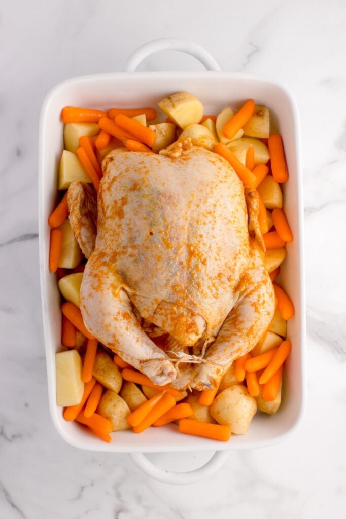 The seasoned raw chicken placed on top of the bed of chopped potatoes and baby carrots inside the white rectangular baking dish, tied with kitchen twine.