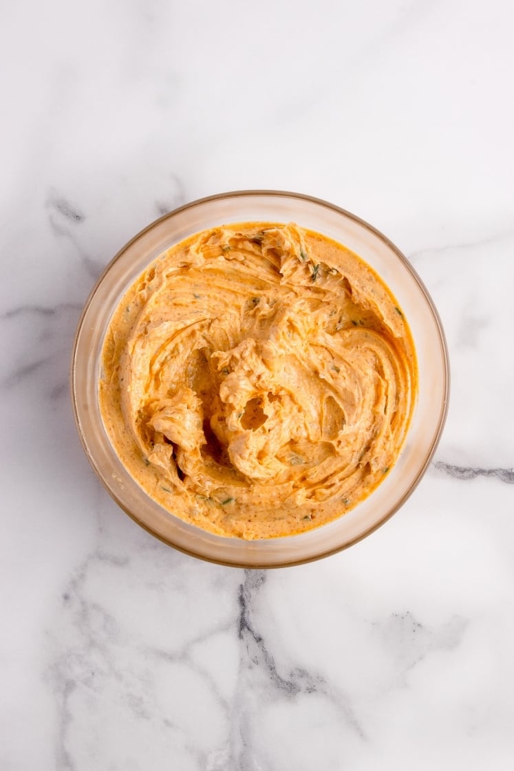 Top-down shot of a glass bowl filled with thick, creamy orange compound butter mixed with chopped green herbs.
