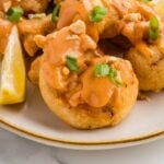Close-up of crispy fried boom boom shrimp coated in creamy orange sauce with green onions, chopped peanuts, and a lemon wedge on a white platter.