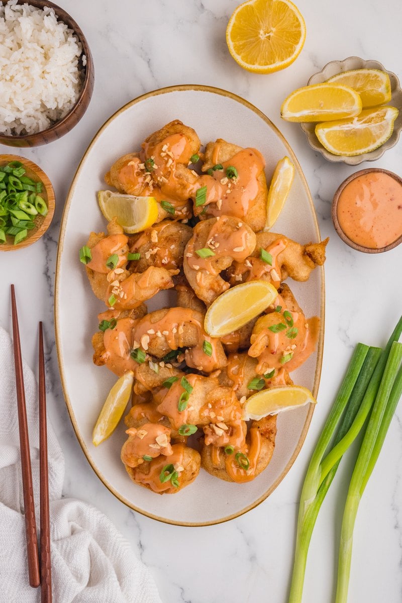 Platter of boom boom shrimp topped with green onions and peanuts with lemon wedges, a bowl of sauce, and chopsticks alongside.