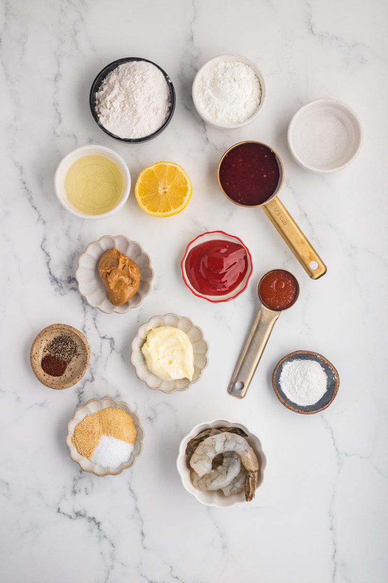 Overhead view of boom boom shrimp ingredients on a marble counter: raw shrimp, flour, cornstarch, baking powder, mayonnaise, sweet chili sauce, ketchup, peanut butter, sriracha, lemon, and spices.
