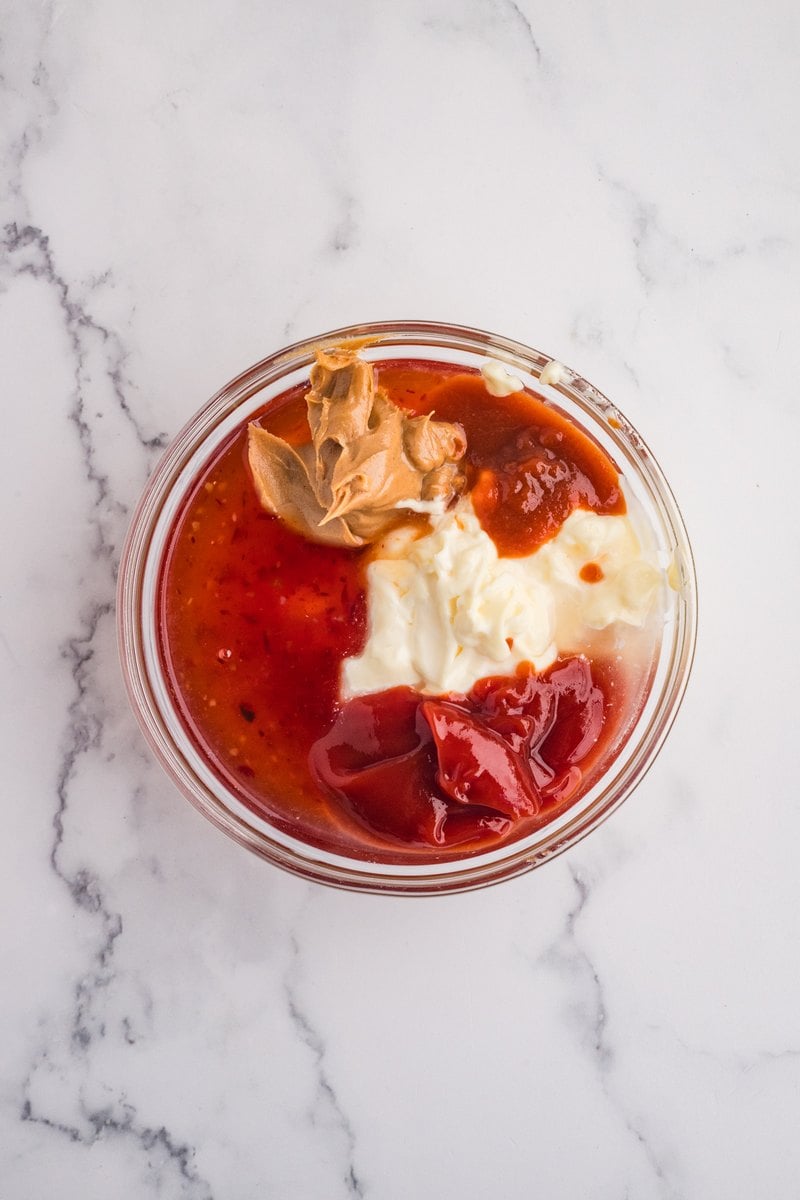 Mayonnaise, sweet chili sauce, ketchup, peanut butter, lemon juice, and sriracha in a glass bowl before whisking.