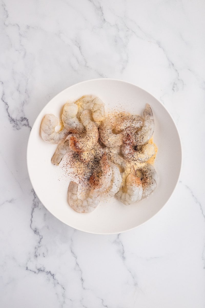Peeled raw shrimp on a white plate sprinkled with salt, garlic powder, pepper, and chili powder.