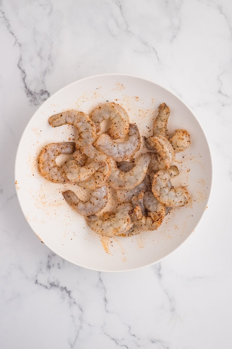 Raw shrimp tossed and evenly coated in the dry seasoning on a white plate.
