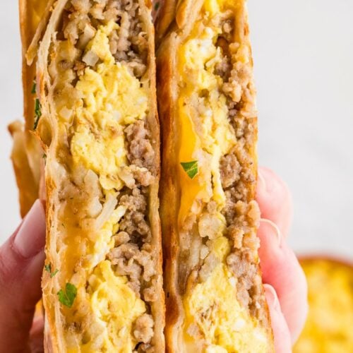 Hand holding two halves of a breakfast crunch wrap showing layers of scrambled egg, sausage, hash browns, and melted cheese inside a golden pan-fried tortilla.