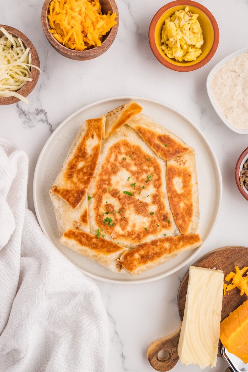 Golden breakfast crunch wrap on a white plate with small bowls of shredded cheese, scrambled eggs, and dipping sauce nearby.