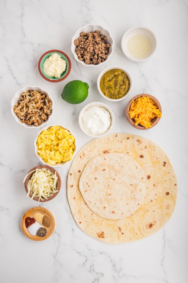 Overhead view of breakfast crunch wrap ingredients on a marble counter: flour tortillas, cooked sausage, scrambled eggs, hash browns, shredded cheddar and Monterey jack, sour cream, salsa verde, mayonnaise, a lime, and spices.
