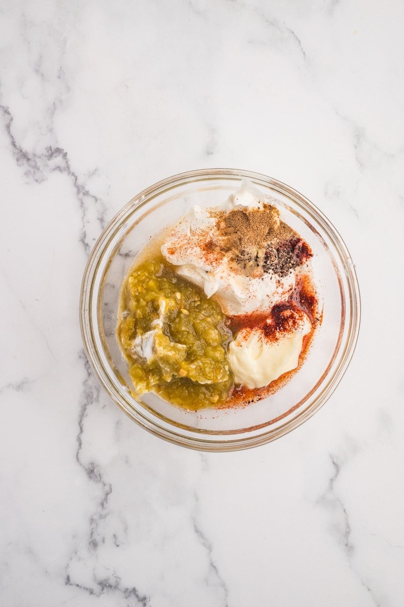 Sour cream, salsa verde, mayonnaise, lime juice, and spices in a glass bowl before whisking.