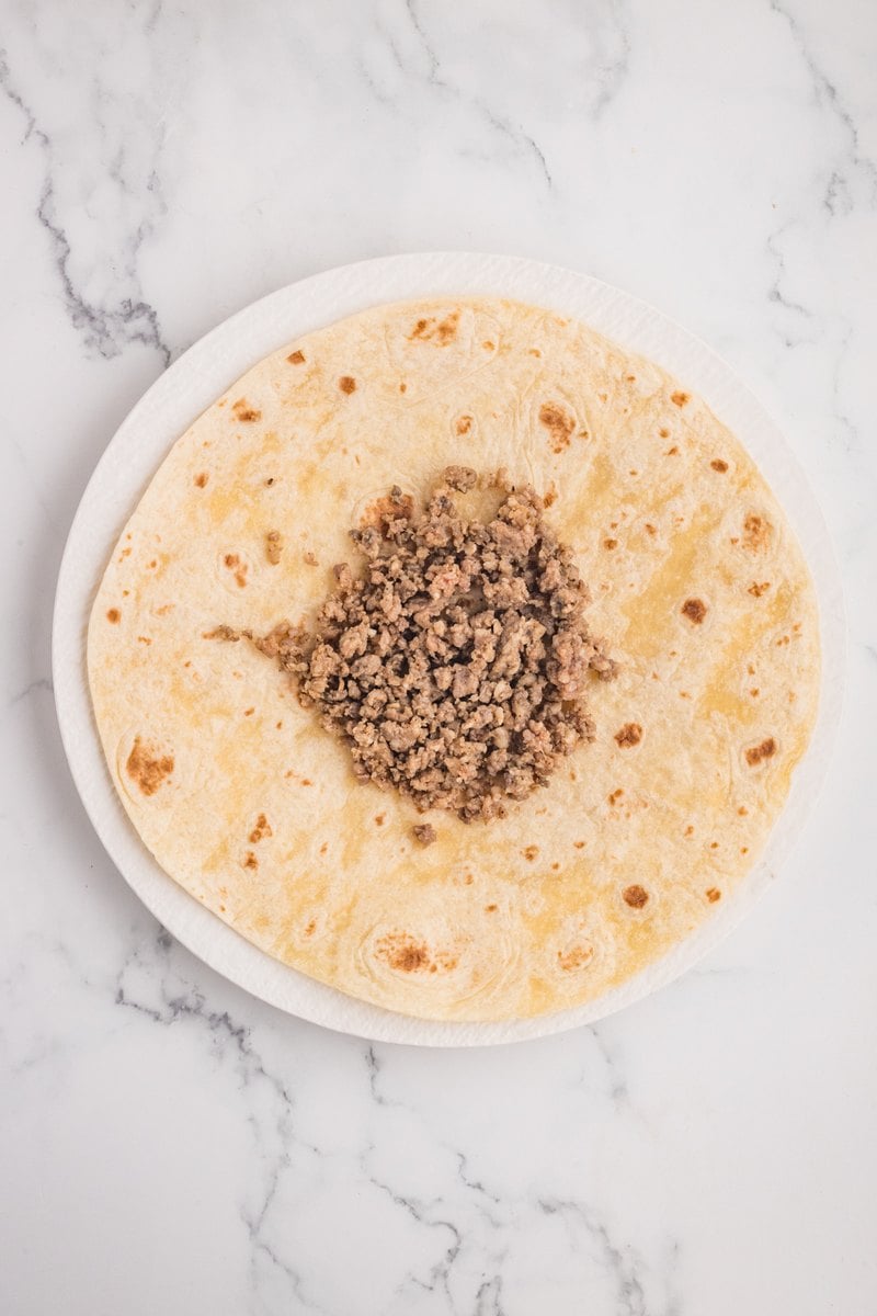 Crumbled cooked breakfast sausage piled in the center of a large flour tortilla on a white plate.
