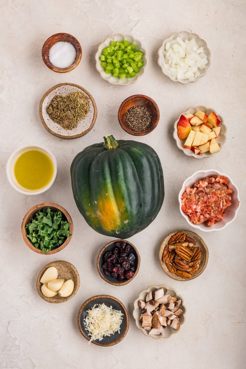 Overhead view of stuffed acorn squash ingredients: a whole acorn squash, ground Italian sausage, diced apple, chopped kale, mushrooms, celery, onion, garlic, dried cranberries, pecans, Parmesan, olive oil, and dried herbs.