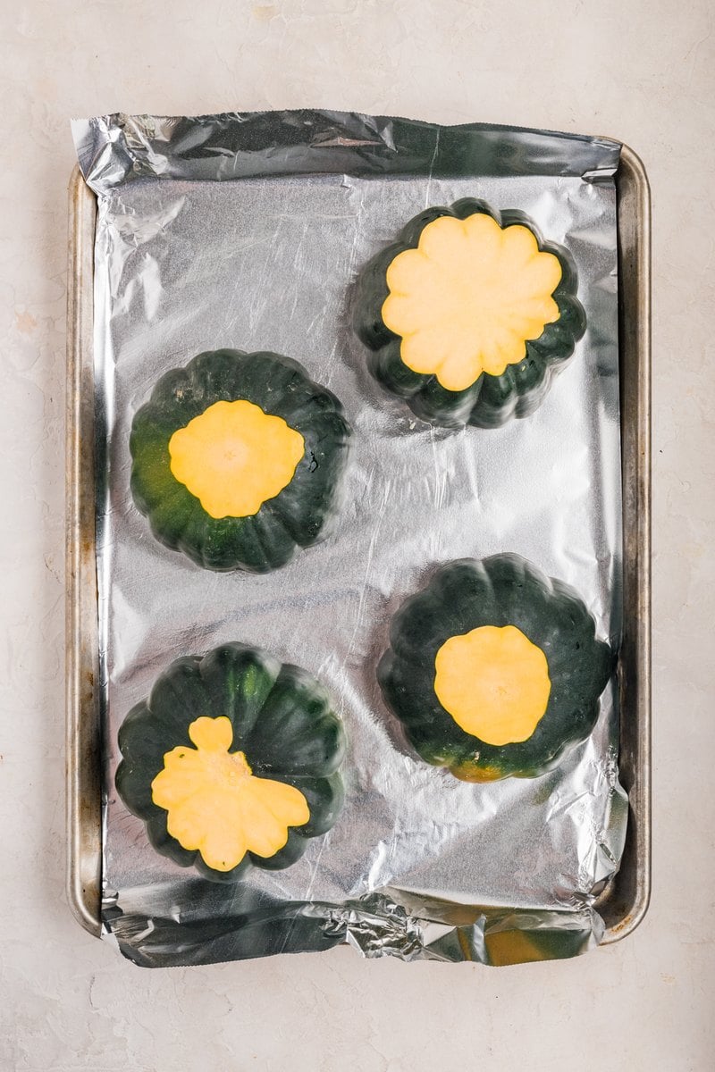Four acorn squash halves cut side down on a foil-lined baking sheet before roasting.