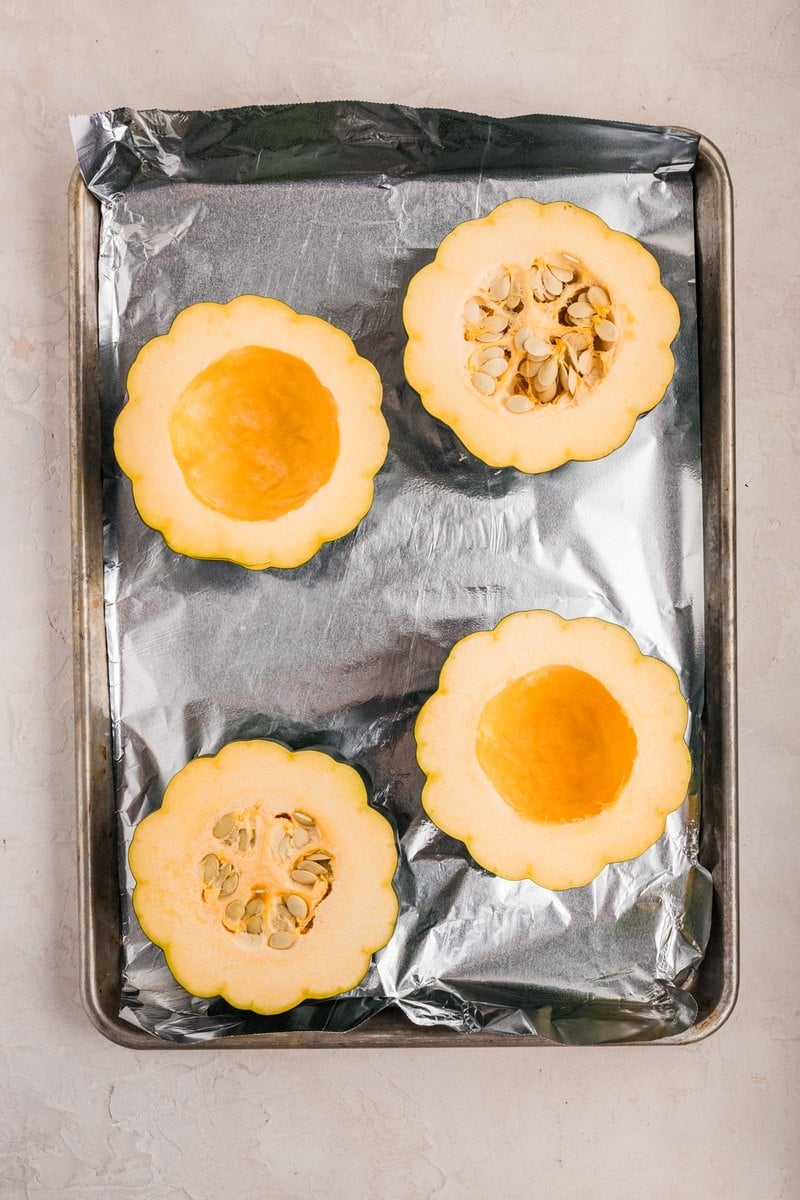 Acorn squash halves with the seeds being scooped out on a baking sheet.